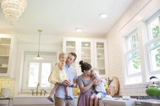 Family in brightly lit kitchen; dad is holding young daughter, mom kissing young son who's sitting on the counter