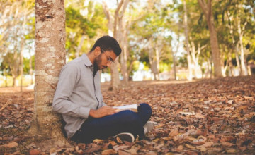 Young man sitting on the ground at the base of a tree studying the Bible