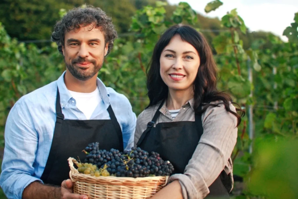 Smiling couple holding a basket of grapes in a vineyard, honoring God through stewardship of the land.