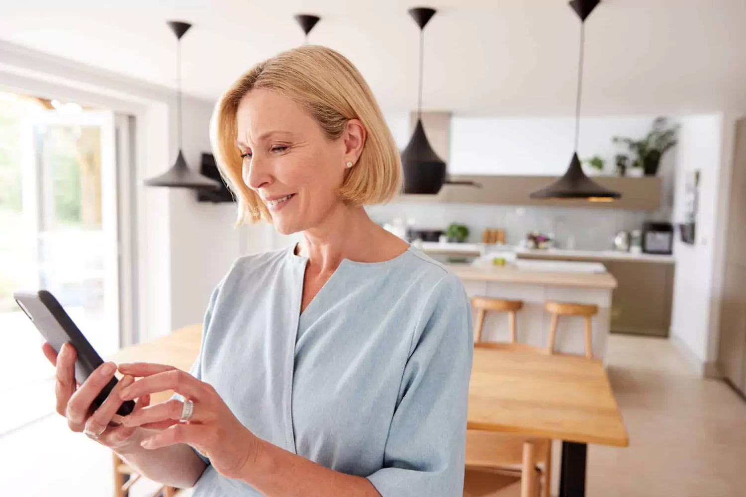 A woman types on her smartphone in a bright kitchen, illustrating how to comfort someone over text with a calm and thoughtful expression.