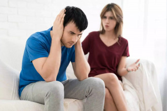 A frustrated man sits on a couch covering his ears while a woman, appearing upset, gestures at him — illustrating the feeling of a husband saying, “My wife constantly criticizes me.”