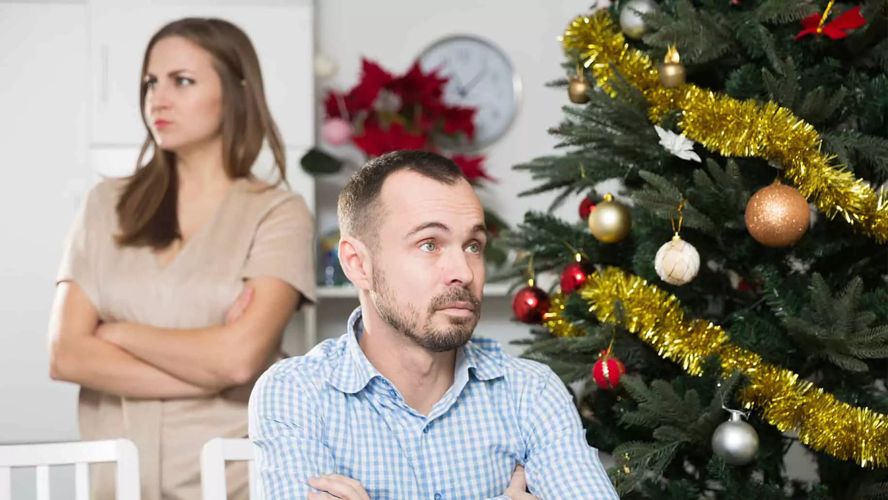 A man and a woman are in a tense moment, displaying signs of holiday stress. The man sits with arms crossed, looking frustrated, while the woman stands behind him, also with arms crossed and an upset expression. A decorated Christmas tree with ornaments and tinsel is visible in the background, highlighting the holiday season amid the tension.