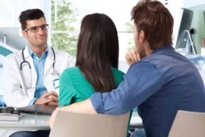 A couple is consulting with a doctor in an office, discussing their efforts to get pregnant. The doctor, wearing a white coat and glasses, listens attentively. The couple sits closely together, indicating support and partnership as they explore the do's and don'ts of trying to get pregnant.