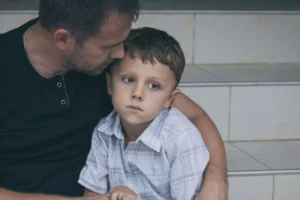 A father is protecting, talking to. and comforting his young son by holding him close on the steps. Protecting and overprotecting your child are different actions.