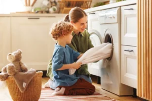 Mom helps child build confidence by teaching him how to do laundry. The image has a little boy and his mom kneeling on the floor talking laundry out of the dryer together