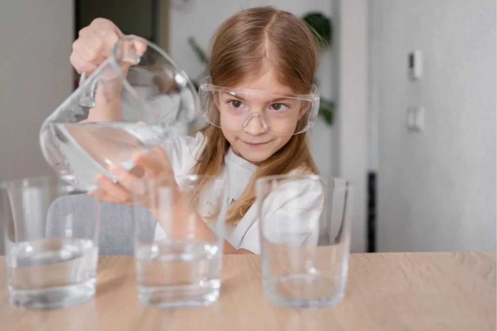 homeschooling little girl doing a science project by pouring water into three glasses wearing plastic goggles