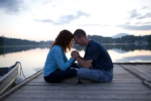 A woman and a man sit on a dock at sunset, holding each other's hands in prayer. Here's how to make God's presence the center of your marriage.