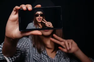 TicTok alternatives. Young teen holding up a phone with herself in the screen holding up a peace sign