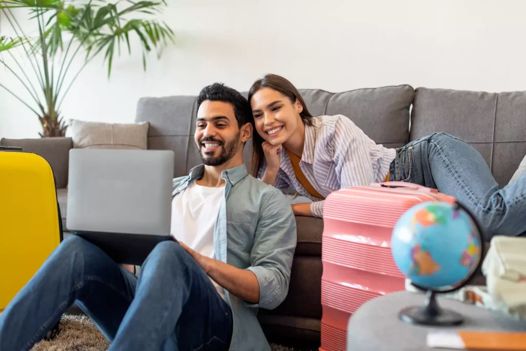 A man sits on the floor with a computer in his lap while his wife lies on the couch looking over his shoulder. Planning your vacation together can help strengthen your marriage.