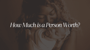 An image of a mother holding her child while the child sleeps with the larger caption in white that says: "How Much Is A Person Worth?"