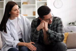 A woman sits next to her dejected husband on a couch, comforting him. Here's how to cope when your spouse is unemployed.