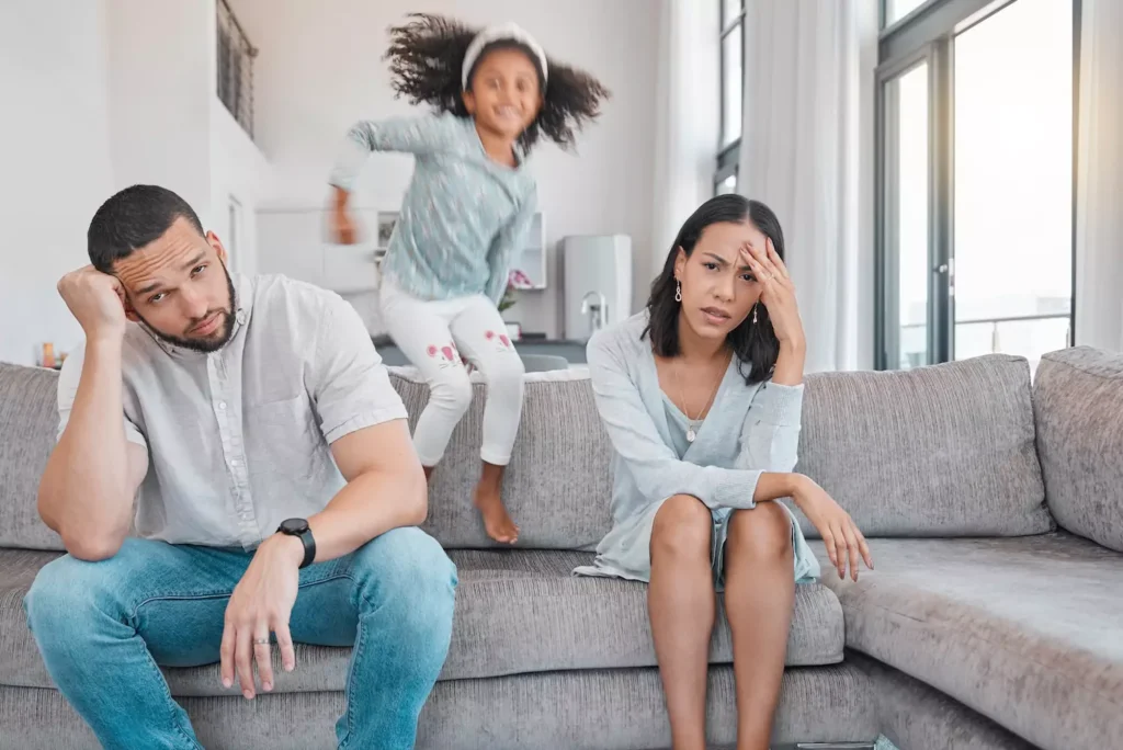 Photo of a man and woman sitting on a couch with their heads in their hands, clearly overwhelmed by their child, who is bouncing on the couch behind them. How you manage parenting stress in marriage can either harm your relationship or create deeper intimacy and happiness.