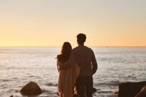 A man and a woman stand arm-in-arm by the seaside at sunset. Life has a way of chipping away at our marriages. Here are a few simple methods to keep romance alive for a successful marriage.