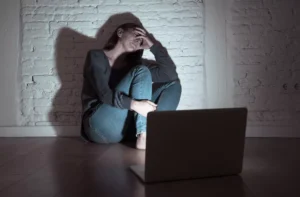 A woman sits in a dark hallway covering her face in shame in the glow of a computer screen. Wives addicted to pornography are not alone.