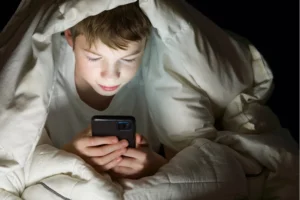 Tech trends sends kids into screen addiction. Boy hiding under the covers late at night staring at his phone screen