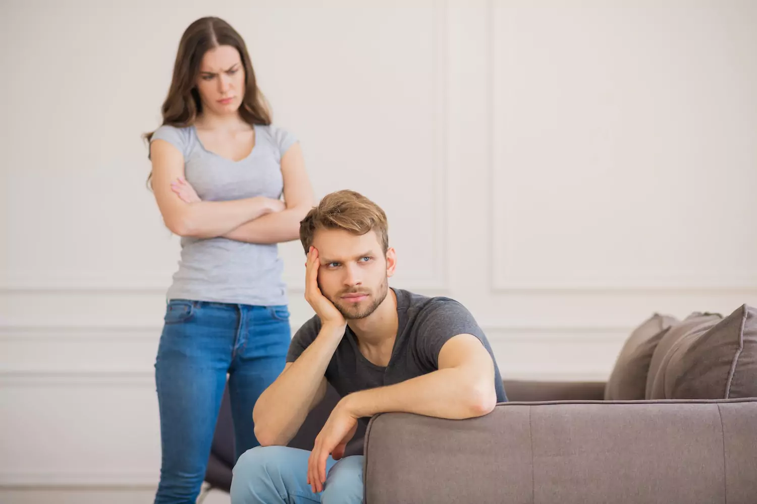 A woman stands, staring angrily at her husband. He sits on the couch, staring off into space in in a detached manner.