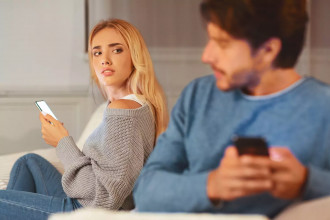 A husband and wife site on a couch. The man is looking over his shoulder at his wife, trying to hide his phone screen from her as she eyes him suspiciously. Here are some warning signs of an extramarital affair and the factors that can lead to them. Take steps today to guard your marriage.