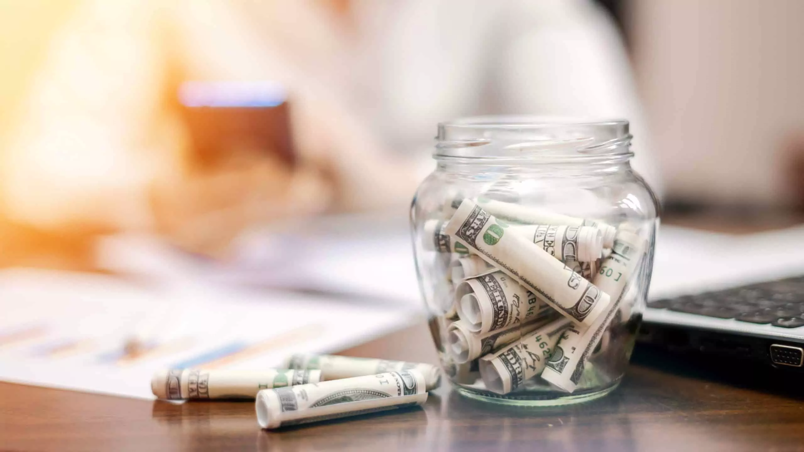 A glass jar filled with rolled-up dollar bills sits on a wooden table alongside a laptop and paperwork. In the background, a person is slightly blurred, suggesting they are working or managing finances after a loss.