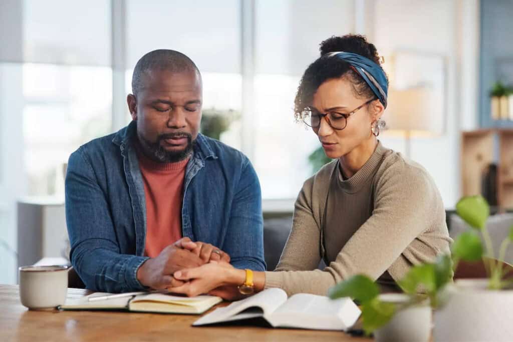 A couple sits at a table holding hands with their eyes closed, praying together in marriage. Books are open in front of them, suggesting a moment of reflection and connection through faith.