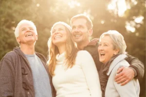 married couples mentors and mentees stand on a rooftop, laughing and enjoying each other’s company on a sunny day. They are casually dressed in colorful sweaters, with arms around each other, and the backdrop features a wooded area