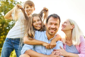 A playful family moment outdoors, showing a father with his daughter on his back, her brother pulling her hair playfully, and the mother smiling affectionately.