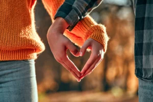 This warm, romantic image captures a couple standing side by side, their hands joining to create a heart shape. The golden hues of autumn leaves in the background add a cozy, intimate atmosphere. The contrast between the textured orange sweater and the plaid shirt enhances the rustic, heartfelt feel of the moment, making it a perfect representation of love, unity, and togetherness.